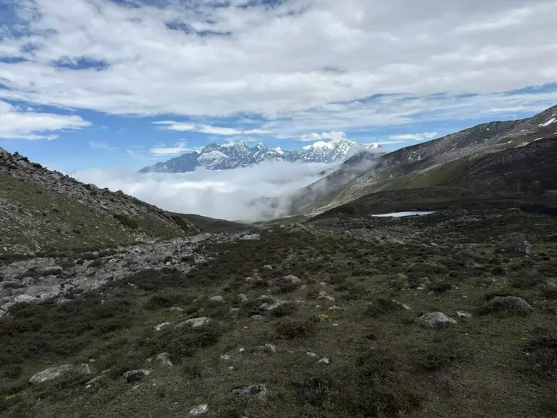 Rocky high-altitude trail cresting above a sea of clouds, with snow-capped peaks in the distance