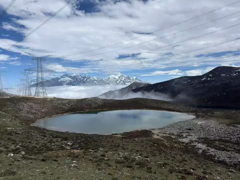 Alpine lake below snow-capped peaks and power transmission lines, with clouds filling the valley below